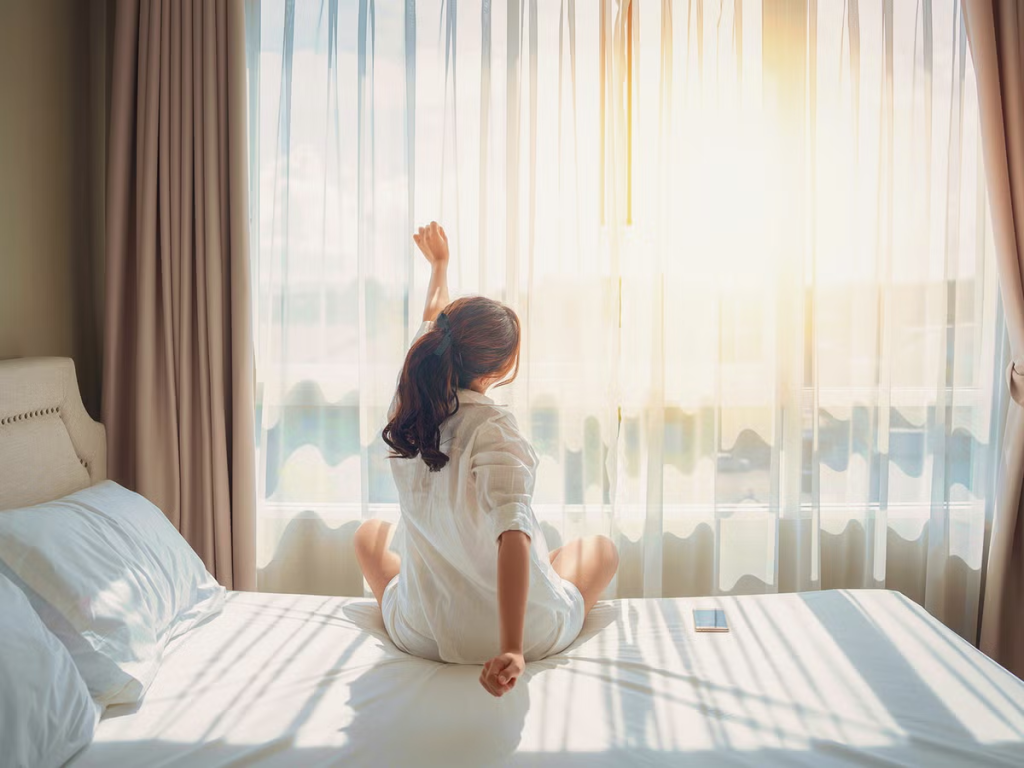 Woman waking up in a comfortable hotel room near Sogo KL, enjoying a relaxing Kuala Lumpur city staycation with natural sunlight.